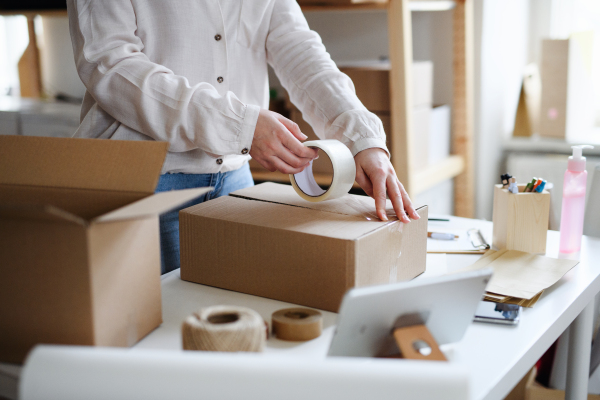 An unrecognizable woman dropshipper working at home, packing parcels. Coronavirus concept.