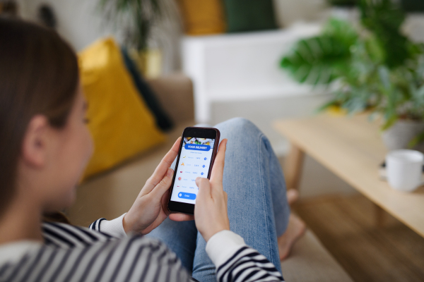 Rear view of young woman with smartphone ordering food at home, coronavirus and food delivery concept.