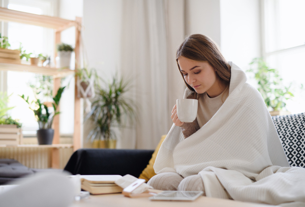 Ill and sick young woman with blanket drinking tea at home, coronavirus concept.