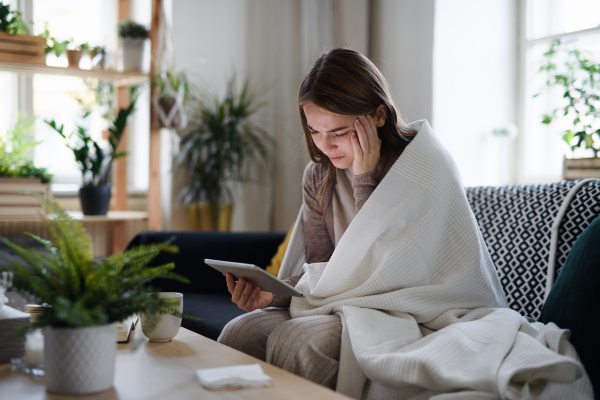 Upset and ill young woman wrapped in blanket on sofa at home, using tablet.