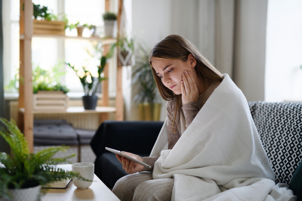 Worried young woman wrapped in blanket using tablet at home, coronavirus concept.