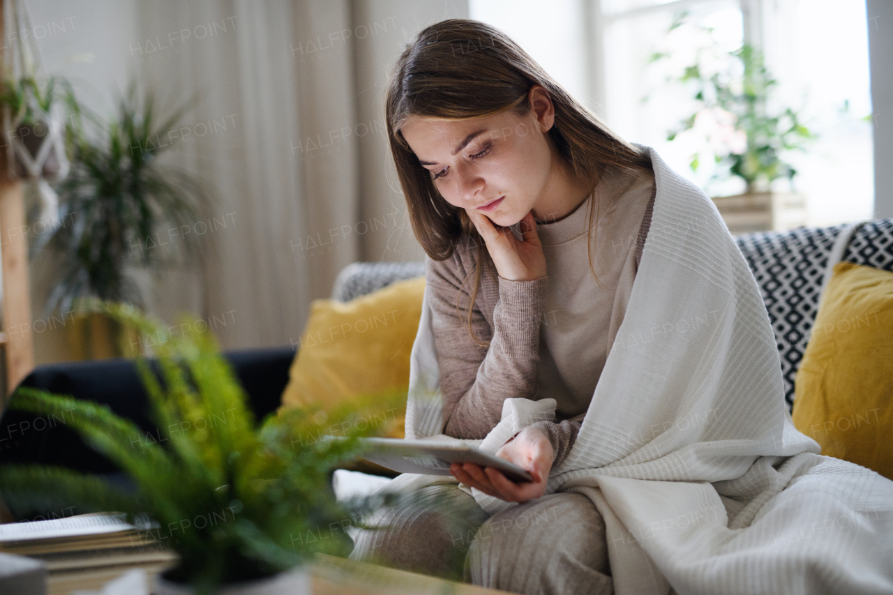 Worried young woman wrapped in blanket using tablet at home, coronavirus concept.