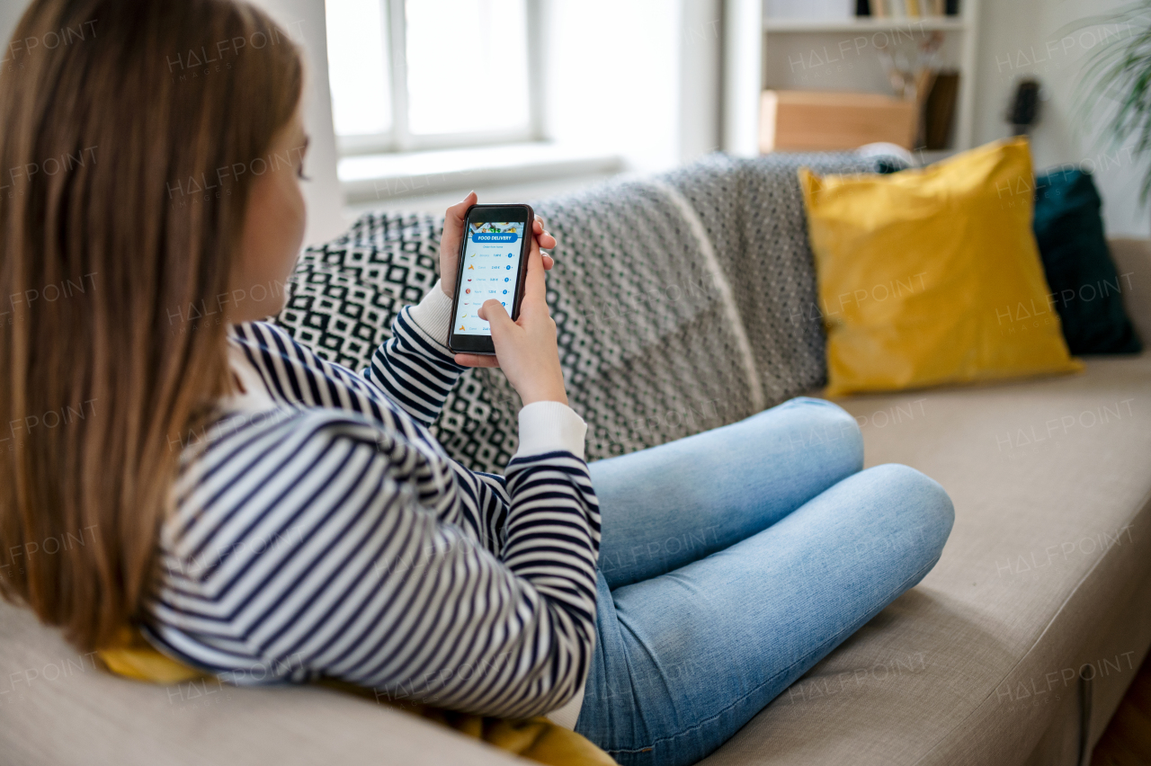 Rear view of young woman with smartphone ordering food at home, coronavirus and food delivery concept.