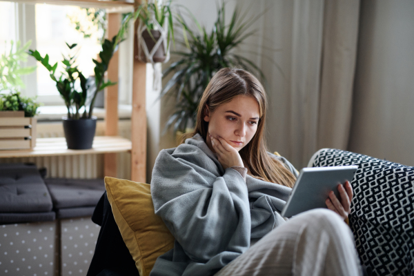 Worried young woman wrapped in blanket using tablet at home, coronavirus concept.