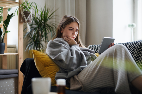 Upset ill young woman wrapped in blanket on sofa at home, using tablet.