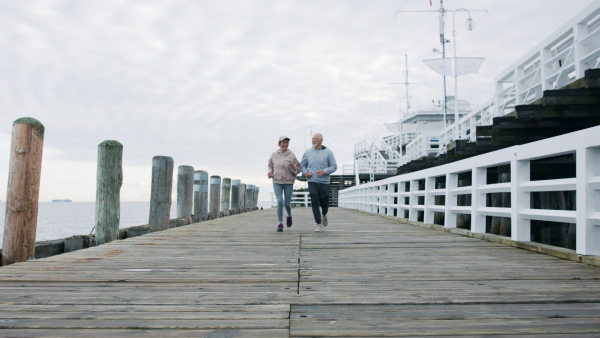 Front view of happy senior couple running outdoors on pier by sea at the morning.