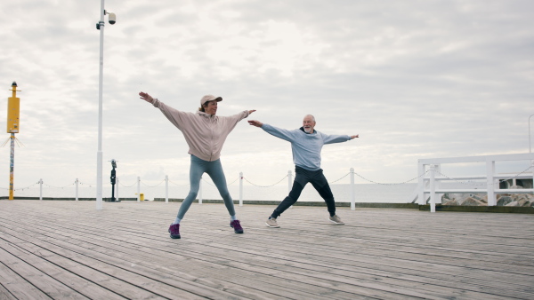 A happy senior couple outdoors on pier by sea at the morning, stretching after run.