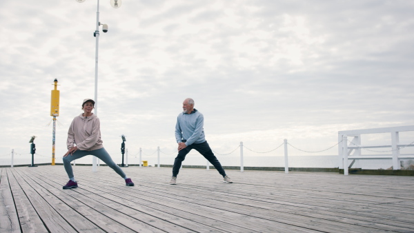 A happy senior couple outdoors on pier by sea at the morning, stretching after run.