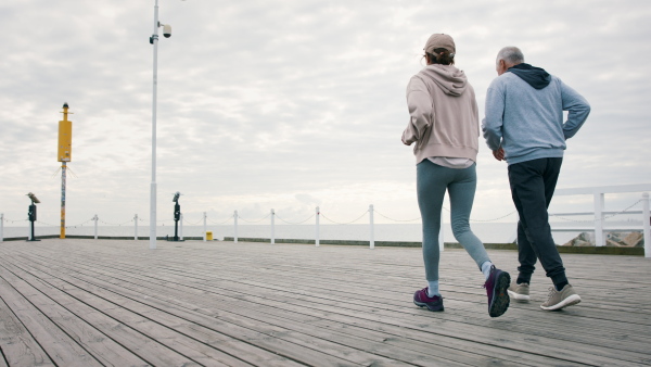 Rear view of happy senior couple running outdoors on pier by sea at the morning.