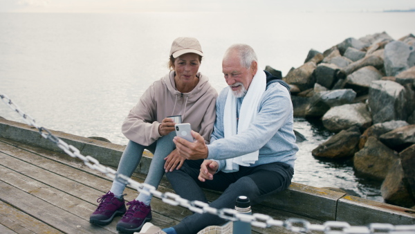 A happy senior couple outdoors on pier by sea at the morning, taking selfie after run.