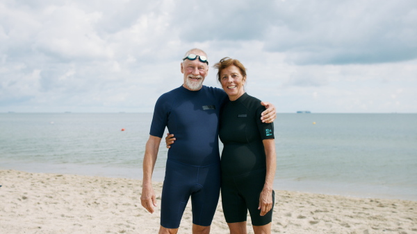 A happy senior couple in neoprene suits outdoors walking on beach at the morning after swimming.