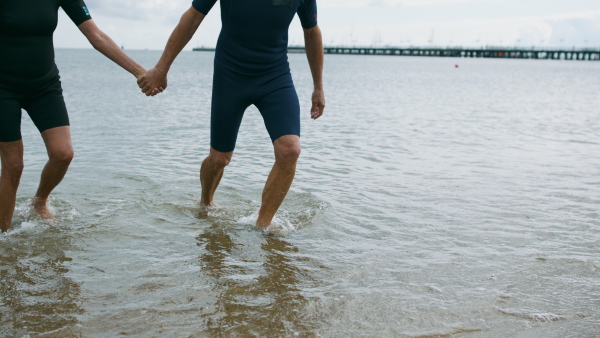 Unrecognizable senior couple in neoprene suits outdoors walking on beach at the morning after swimming.