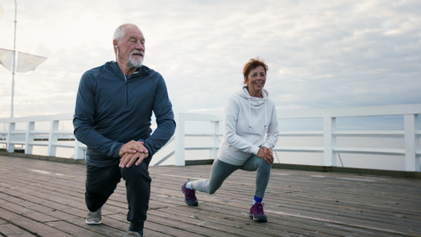 A happy senior couple outdoors on pier by sea at the morning, stretching after run.