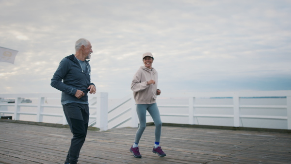 A happy senior couple running outdoors on pier by sea at the morning, hugging.