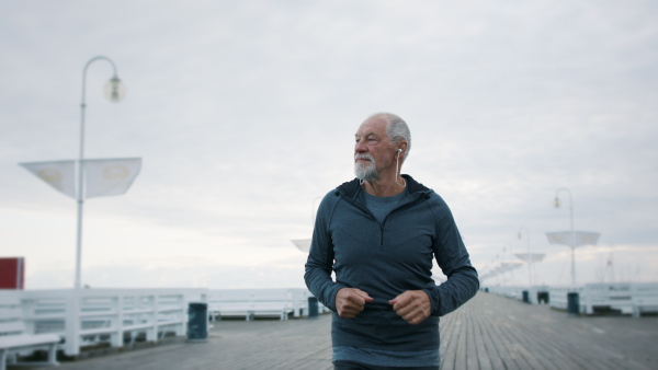 Front view of happy senior man running outdoors on pier by sea at the morning.