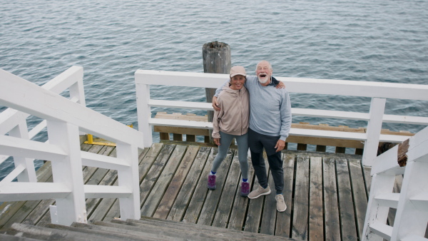 A happy senior couple outdoors on pier by sea at the morning, hugging after run.