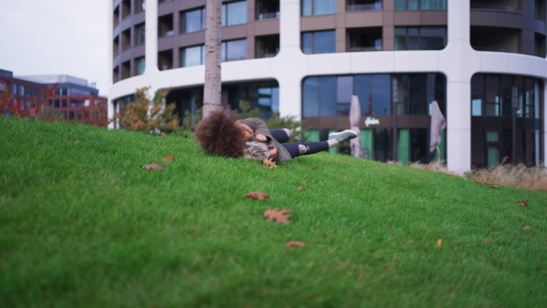 A little Afrcian American girl rolling down grassy hill outdoors in park in city in autumn day.