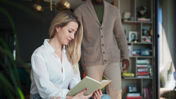 A mid adult caucasian woman reading book when her african american husband comes and kisses her at home.