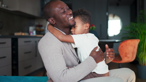 A mid adult african american father sitting and hugging his little son at home, multiracial family concept.