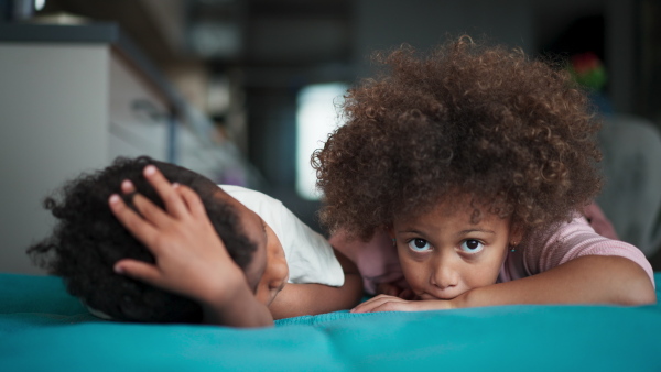 Two little multiracial kids siblings lying on front on a bed and looking at camera at home.