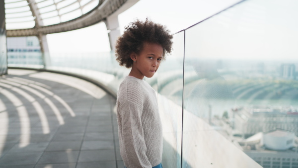 A little multiracial girl with afro hairstyle standing on bridge and looking at city view outdoors.