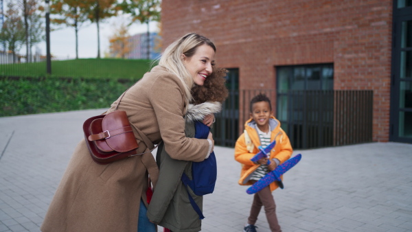 A happy mid adult mother waiting for her little multiracial kids after school outdoors in street, hugging and kissing them.