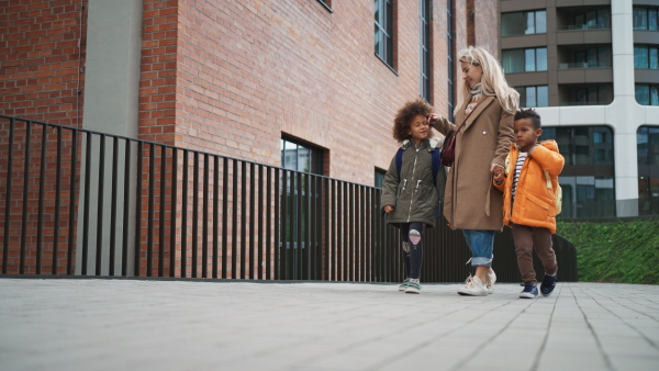 A low angle view of happy mother taking children home from school, walking outdoors in street, multiracial family.