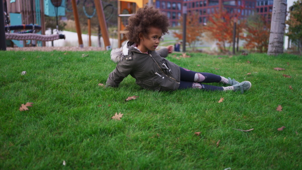 A little Afrcian American girl rolling down grassy hill outdoors in park in city in autumn day.