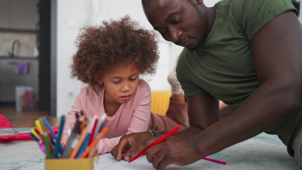 A little multiracial girl lying on floor and doing homework with his african american father at home.