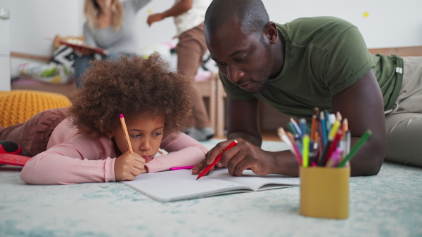 A little multiracial girl lying on floor and doing homework with his african american father at home.