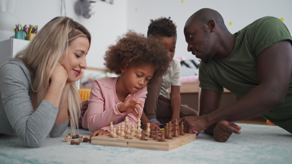 A young multiracial family with two little kids lying on floor and playing chess at home.