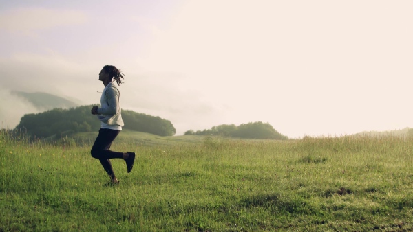 Side view of young mixed race man with dreadlocks running on meadow outdoors in nature.