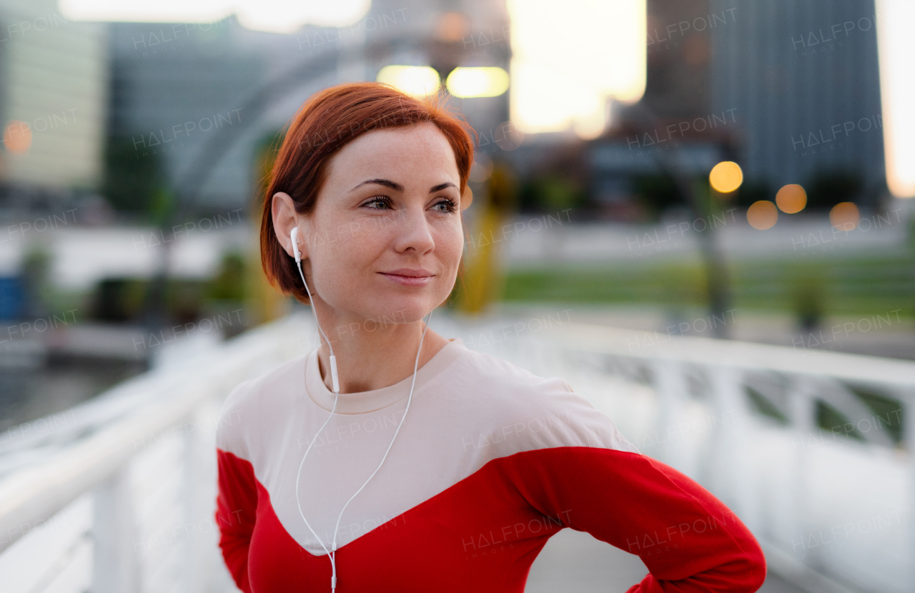 Front view of young woman runner with earphones in city, resting on the bridge.