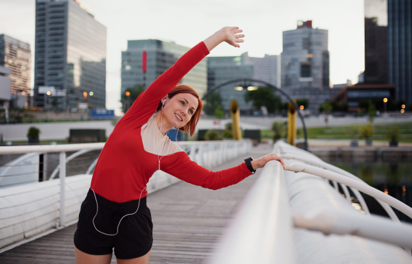 Front view of young woman runner with earphones in city, stretching on the bridge.