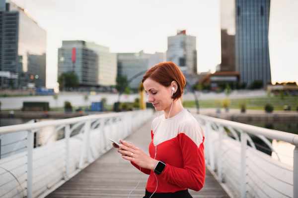 Young woman runner with earphones in city, using smartphone when resting.