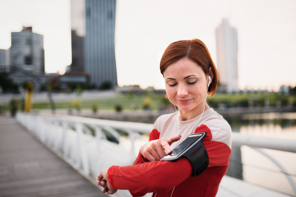 Young woman runner with earphones in city, using smartphone when resting.