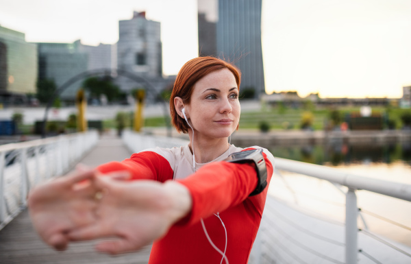 Front view of young woman runner with earphones in city, stretching on the bridge.