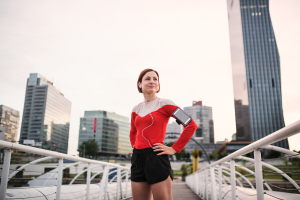 A young woman with earphones and smartphone standing outdoors in city, resting after exercise.