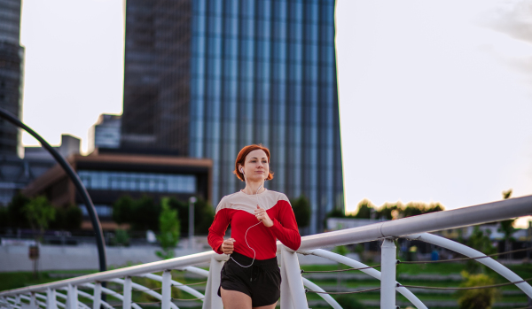 Front view of young woman runner with earphones jogging outdoors in city.