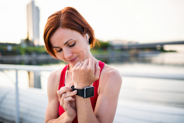 Young woman runner with earphones in city, using smartwatch when resting.