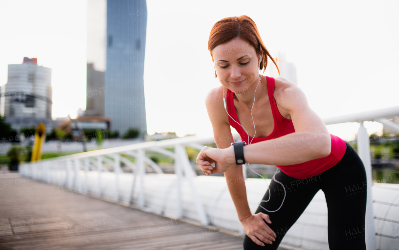 Young woman runner with earphones in city, using smartwatch when resting.