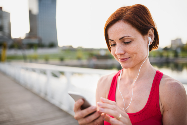 Young woman runner with earphones in city, using smartphone when resting.