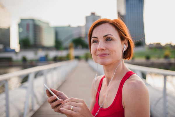 Young woman runner with earphones in city, using smartphone when resting.