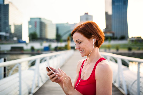 Young woman runner with earphones in city, using smartphone when resting.