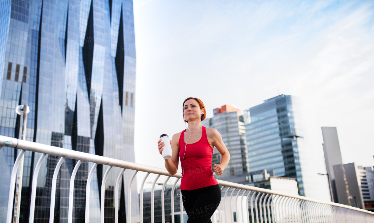 Active young woman runner with earphones and water bottle jogging outdoors in city.