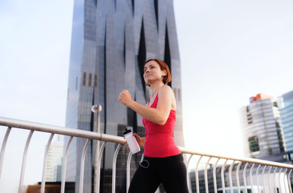 A young woman with earphones doing exercise on bridge outdoors in city, running.