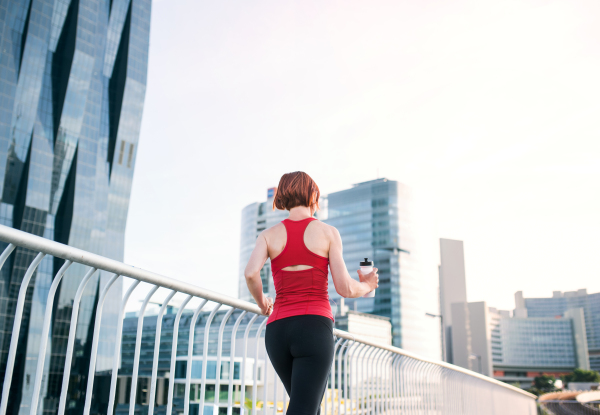 Rear view of active young woman runner with water bottle jogging in city.