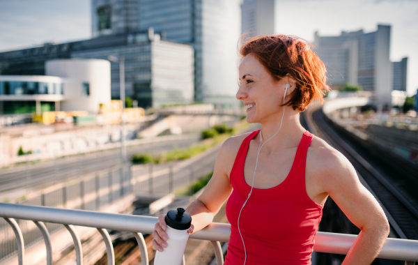 Young woman runner with earphones and water bottle in city, resting on the bridge. Copy space.