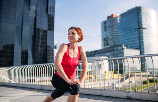 A young woman with earphones doing exercise on bridge outdoors in city, stretching.