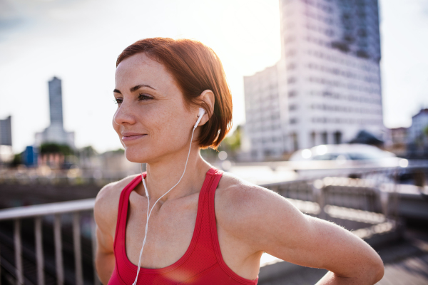 Front view of young woman runner with earphones in city, resting on the bridge.